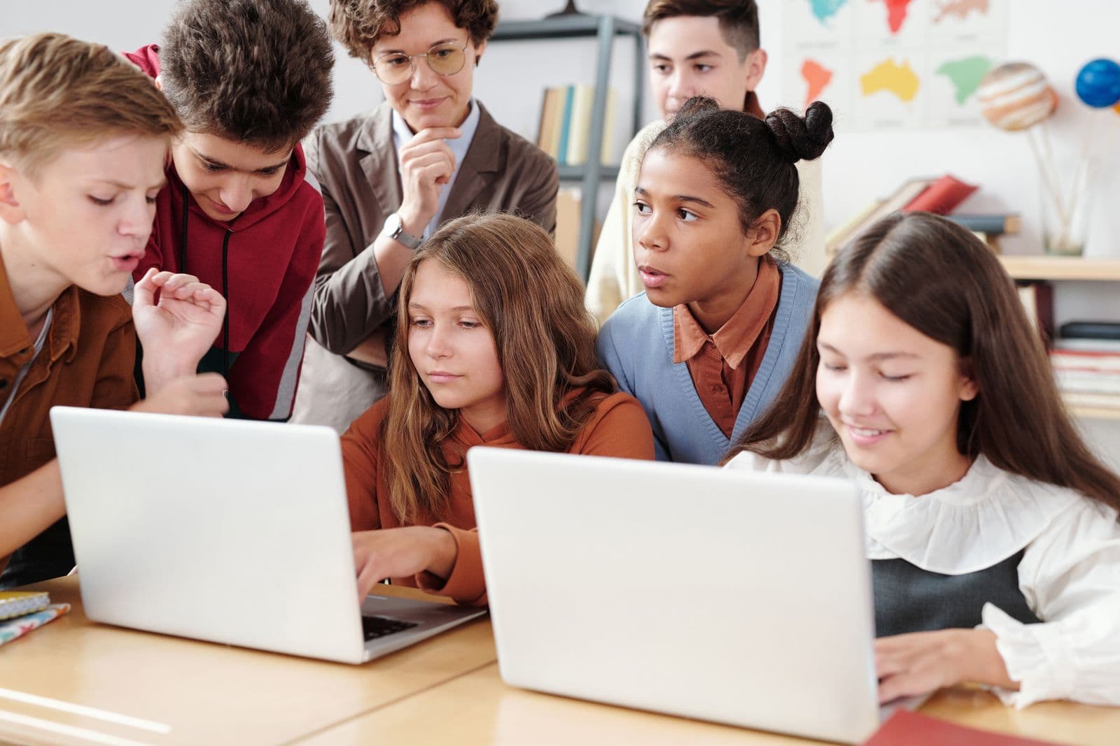 A group of young students crowded around two laptops in a classroom while a teacher looks on.