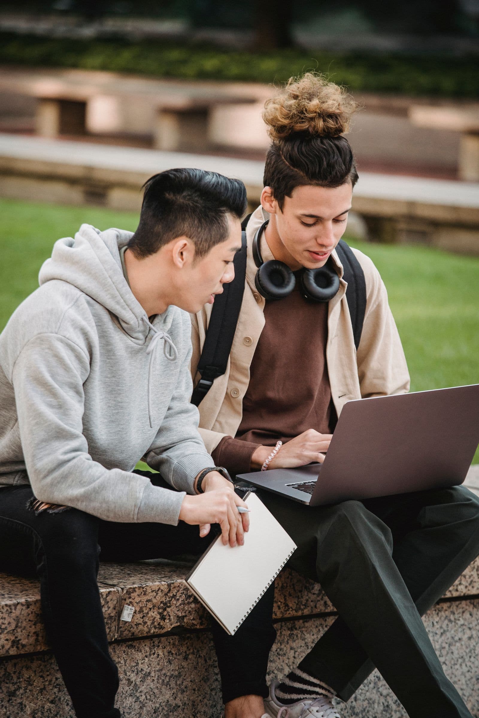 Two young men sitting on a stone bench outdoors, one typing on a laptop while the other holds a notebook.