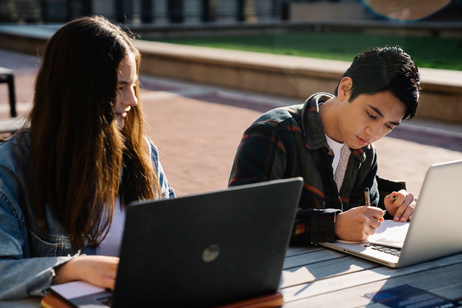 Two young people working on laptops at an outdoor table in afternoon light, one taking notes by hand.
