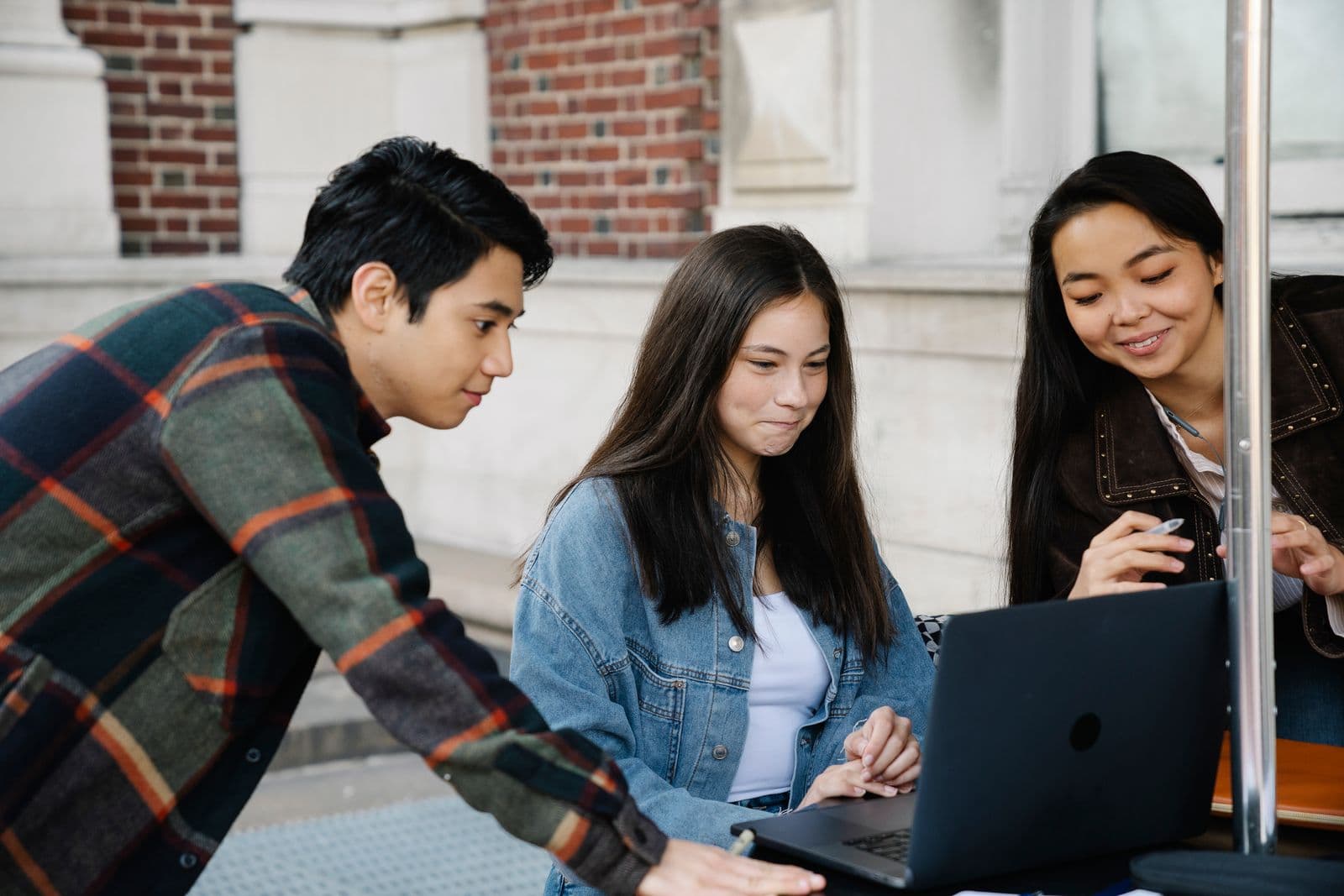 Three young adults gathered around a laptop outside a brick building, smiling at the screen.