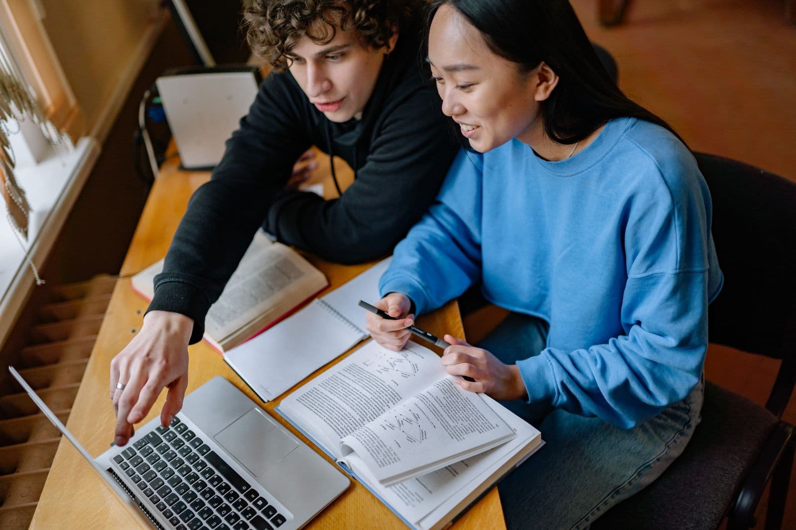 Two students at a wooden table working through a problem together on a laptop and an open textbook.
